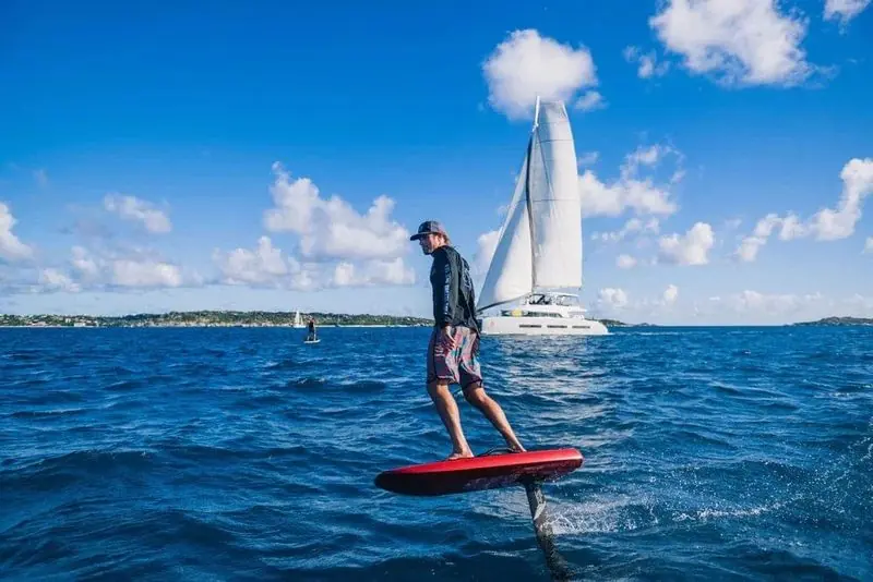 Blue water and a moored catamaran frame a rider standing on a red foil board.