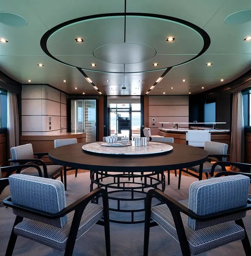 A round dining table in the saloon of a motor yacht, ringed by armchairs upholstered in a blue and white woven cloth under a circular light trough in the deckhead.