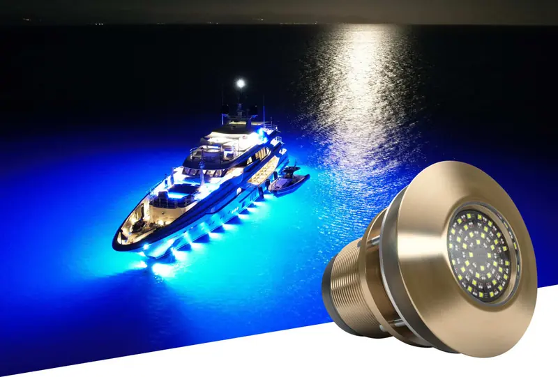 A superyacht anchored at night, its hull ringed in blue Lumishore underwater light spreading across the water, inset with a studio shot of a bronze thru-hull fixture.