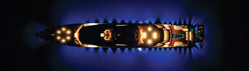 A superyacht seen from directly overhead at night, deck lighting picking out the foredeck and cockpit as the hull cuts through dark water.