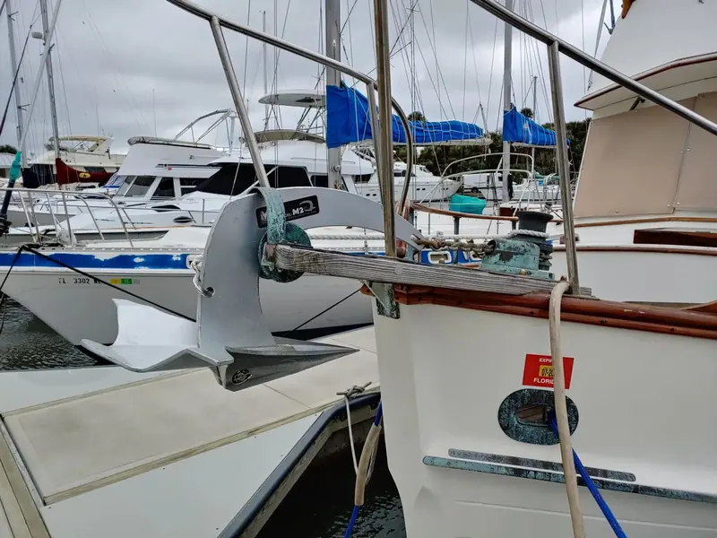 A Mantus M2 anchor stowed on the bow roller of a Grand Banks 36 trawler, the anchor's own M2 branding visible on the shank, with the marina's other liveaboard boats behind it under an overcast Florida sky.