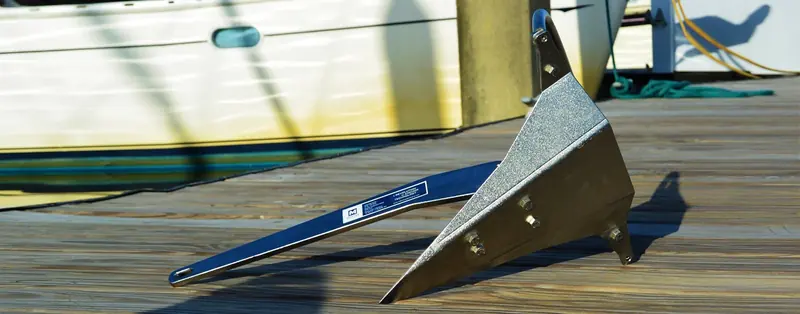 A Mantus anchor resting fluke-down on a sunlit dock, its Revolution in Anchor Technology decal visible on the shank, showing the roll-bar geometry and the galvanized finish in direct light.