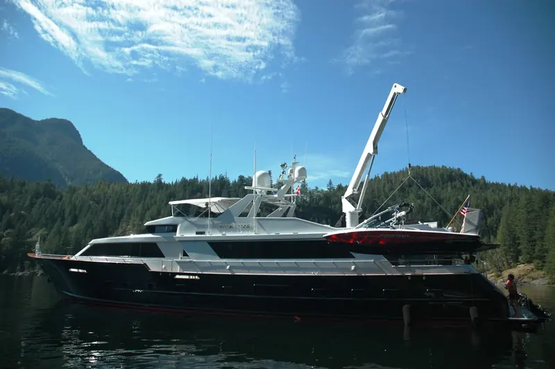 A crane swings a MasterCraft tender out from the stern of the motor yacht Tawnie Lynne, anchored beneath a tree-lined shore.