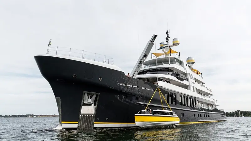 A knuckle-boom crane lowers a yellow limousine tender alongside the black hull of the explorer yacht Scout as two crew guide it down.