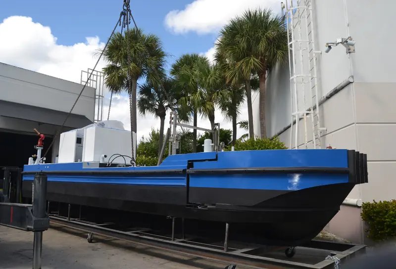 A Scavenger 2000 aquatic weed harvester hull being rigged for lifting at the builder's Florida facility, palm trees behind. Northern Lights generators power the harvester's spray and drive systems.