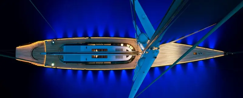 A sailing yacht seen from directly overhead at night, its Ocean LED deck-edge lighting tracing the sheerline in gold against the blue-lit water either side of the hull.