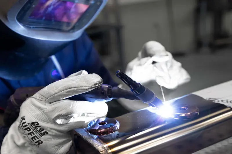 A welder in a leather glove stamped Kupfer runs a TIG torch along a stainless steel platform arm in Opacmare's own workshop, the weld pool bright against the polished tube.