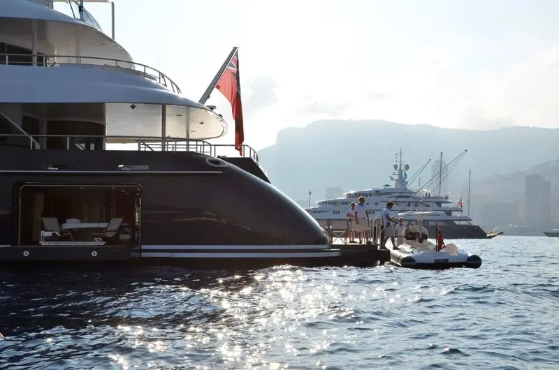 Guests step from a superyacht swim platform into a tender alongside, the beach club open behind them.