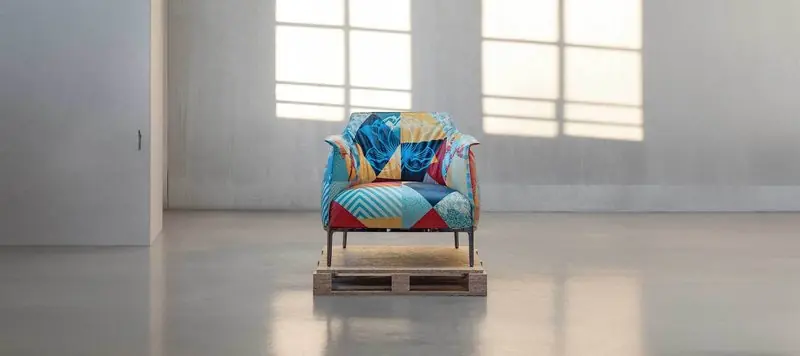 An Archibald armchair upholstered in patchwork art fabric stands on a travertine pallet in a bare gallery space.