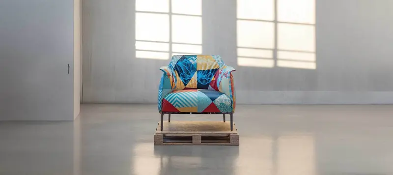 An Archibald armchair upholstered in patchwork art fabric stands on a travertine pallet in a bare gallery space.