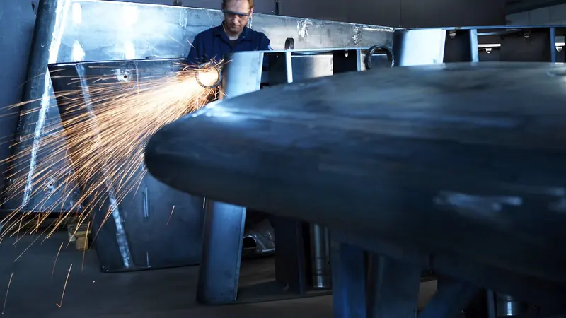 A technician grinds a weld seam on a fin-stabilizer housing during fabrication, the raw casting still in jig.