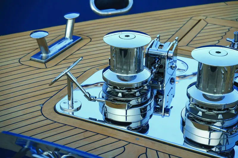 A pair of Quick vertical windlass capstans mounted on polished stainless backing plates set into a teak foredeck, chain visible through the open gypsy housing beside the manual handle socket