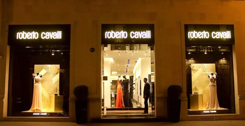 Lit signage names Roberto Cavalli above three night windows holding gowns on mannequins.