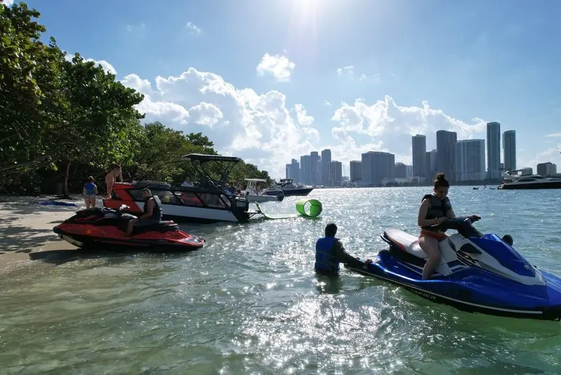 A red Sea-Doo and a Sea-Doo Switch pontoon rest on a sandbar while riders wait in the shallows, the city skyline across the bay.