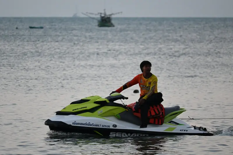 Sitting side-saddle on a green and white Sea-Doo GTI, a rider drifts on flat grey water with a fishing boat behind.