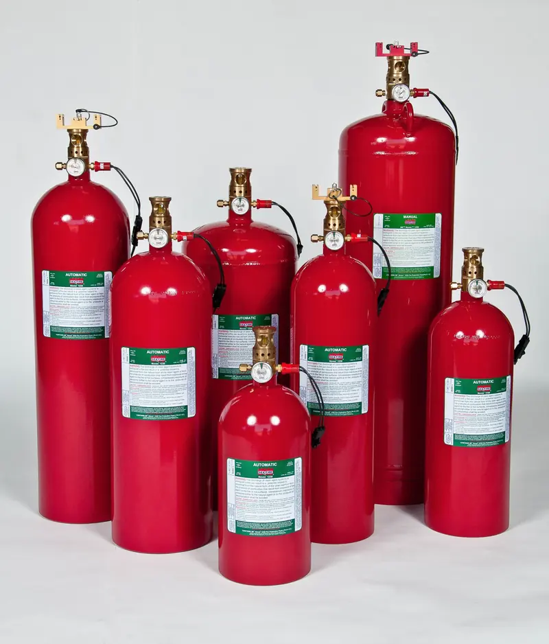 Eight red Sea-Fire cylinders of different heights stand grouped on a studio floor, each topped with a brass valve head and pressure gauge, wired leads coiled beside them.