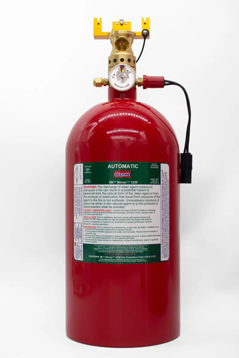 Close on a single red NFD cylinder's brass valve assembly and pressure gauge, the green Sea-Fire label below it printed with USCG approval and Novec 1230 fill details.
