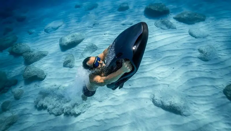 A diver drives a dark Seabob low over a rippled sand bottom, bubbles streaming behind him.