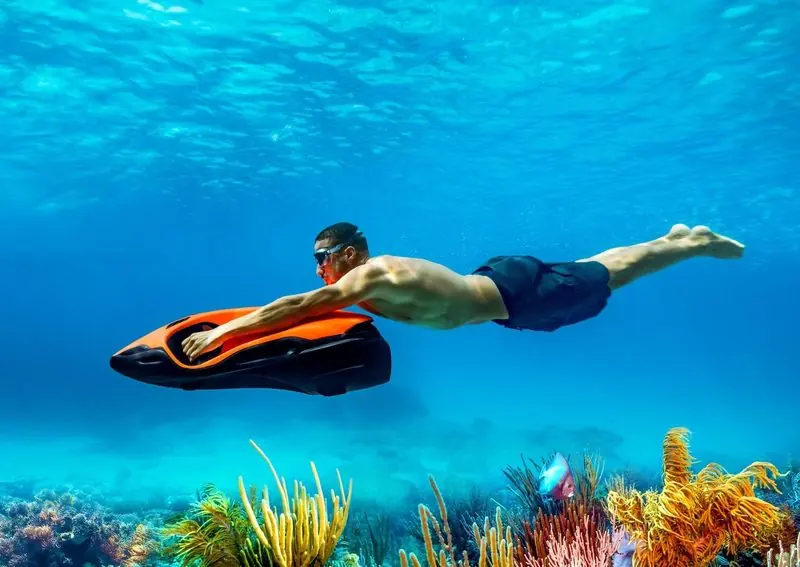 Underwater, a swimmer holds an orange and black Seabob level above a reef of soft corals.