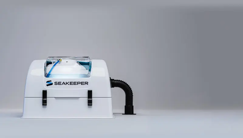 A white Seakeeper stabiliser stands on a studio floor, the clear inspection cover on top showing the enclosed sphere and its blue feed line. A ribbed black duct curves away from one side to a round deck flange.