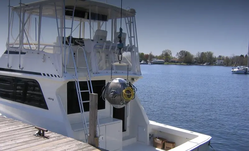 A polished steel gyro sphere hangs from a hoist over the cockpit of a Viking sportfishing boat lying at a timber dock, cabling coiled in loops around it and cardboard parts boxes on the deck below.