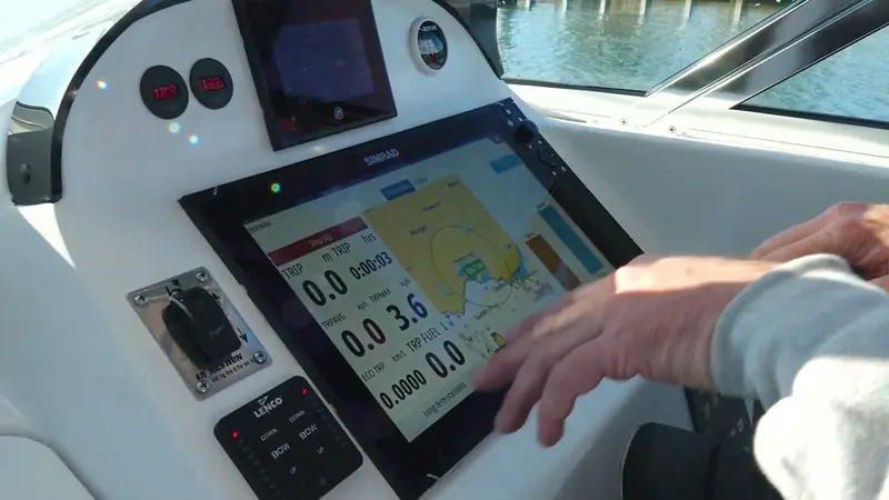 A Simrad multifunction display built into a helm dash alongside a Lenco trim tab control and analog gauges, with a hand operating the touchscreen underway.