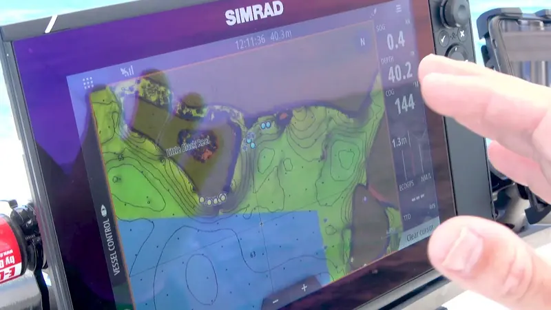 Close-up of a Simrad chartplotter touchscreen showing a contour chart with depth, speed and course data, a finger tapping the vessel-control panel.