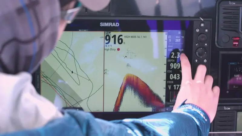 A boater at the helm reading a Simrad multifunction display split between chartplotter and sonar screens while underway.