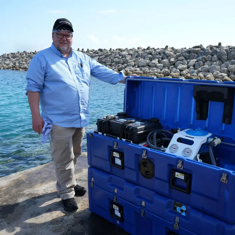 A man in a work shirt stands beside an open blue transport case holding a portable Spectra watermaker system, a rock breakwater and turquoise water behind him