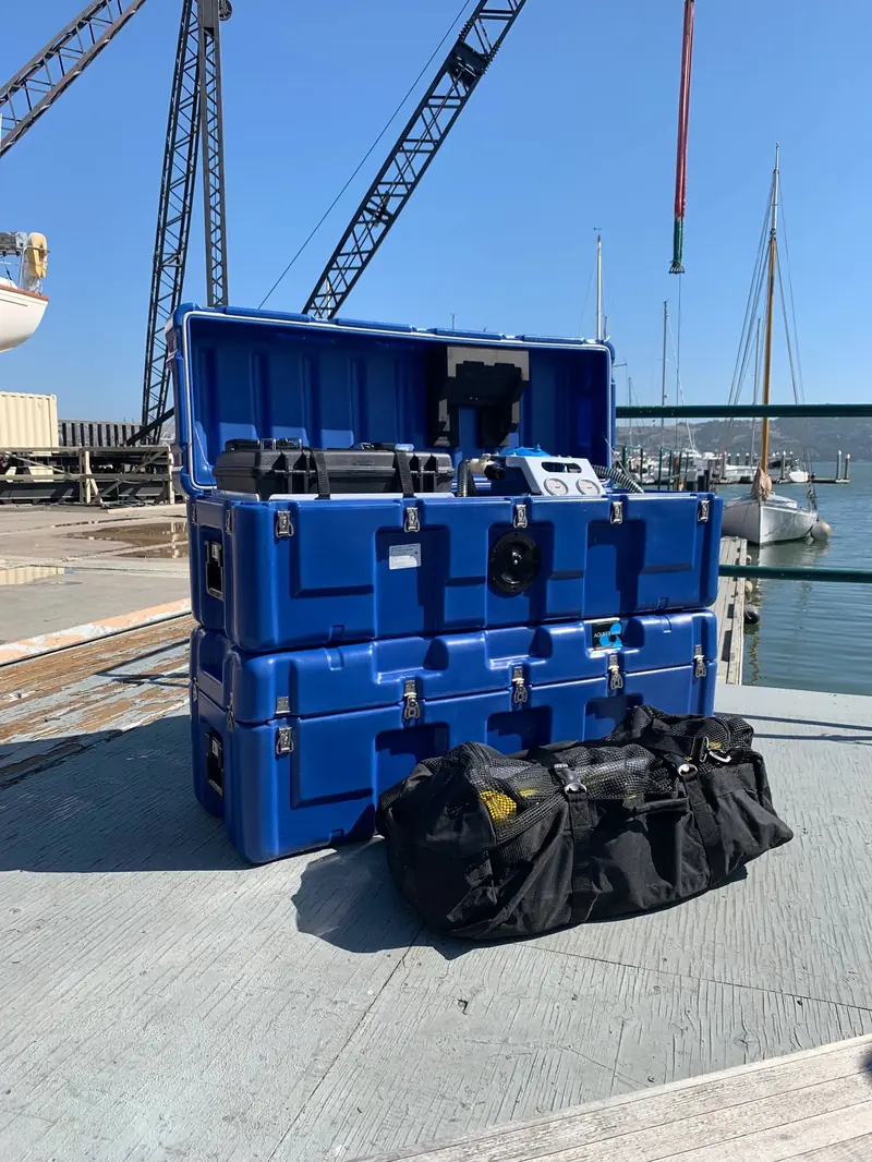 A stacked pair of blue watermaker transport cases sits open on a concrete dock beside a black carry bag, sailboat masts and a harbour crane in the background
