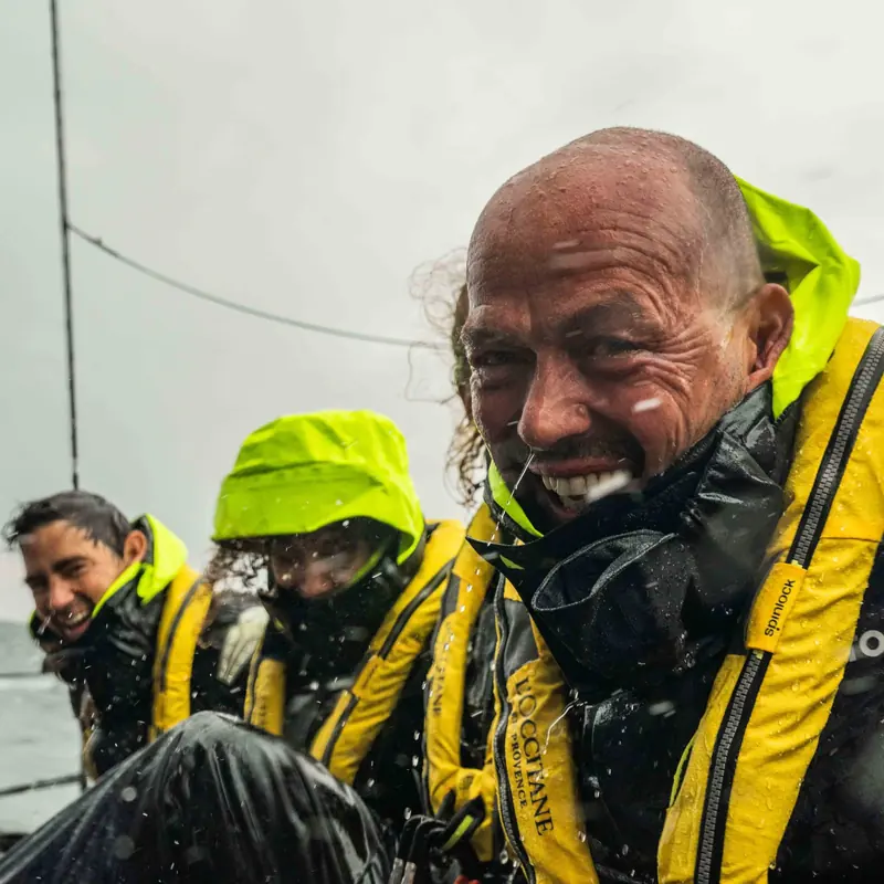 Spray soaks an offshore crew in yellow lifejackets during a wet leg, the nearest wearing a Spinlock branded harness tab.