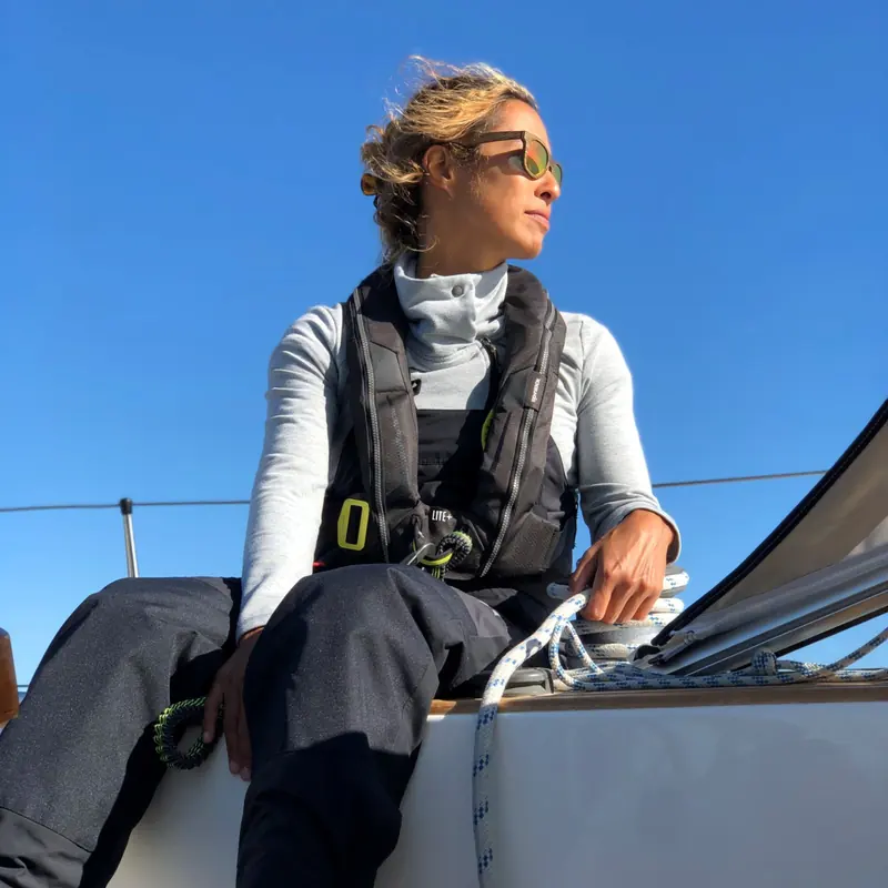 A sailor in a Spinlock deckvest sits on the rail, watching the sail trim as the boat heels under a clear sky.