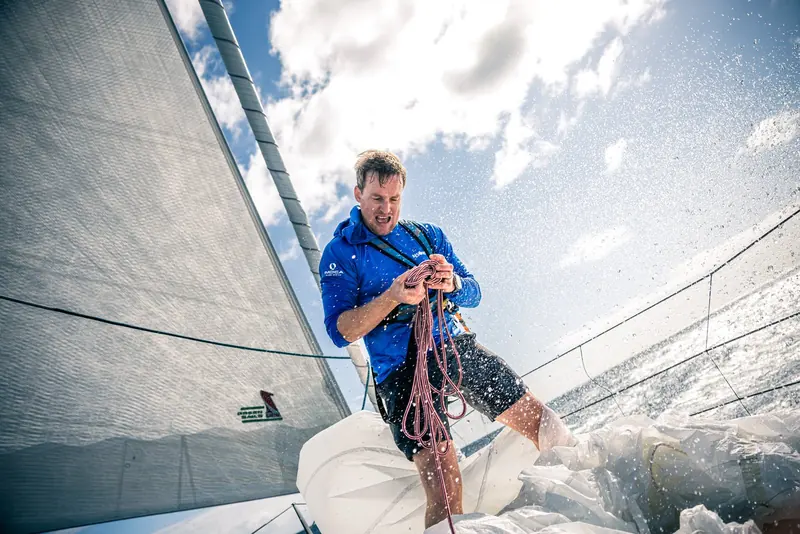 A crewman leans out over spray on the foredeck, gathering a headsail while clipped into a harness line.