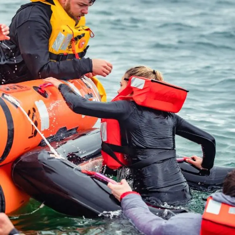 A crew member in immersion gear reaches down to help a swimmer climb aboard an open reversible liferaft during a sea survival drill.