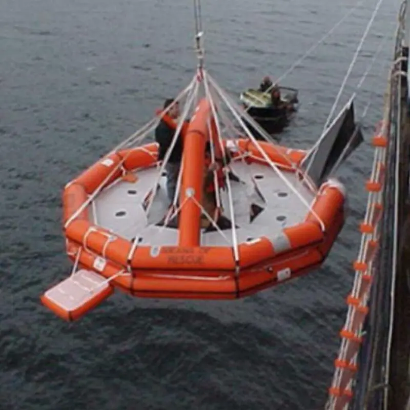 A means-of-rescue liferaft is lifted clear of the water by crane, a support boat standing by during a recovery exercise.
