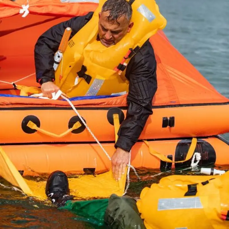 A crew member kneels at the boarding ramp of an inflated leisure liferaft, pulling a person from the water by hand during a drill.