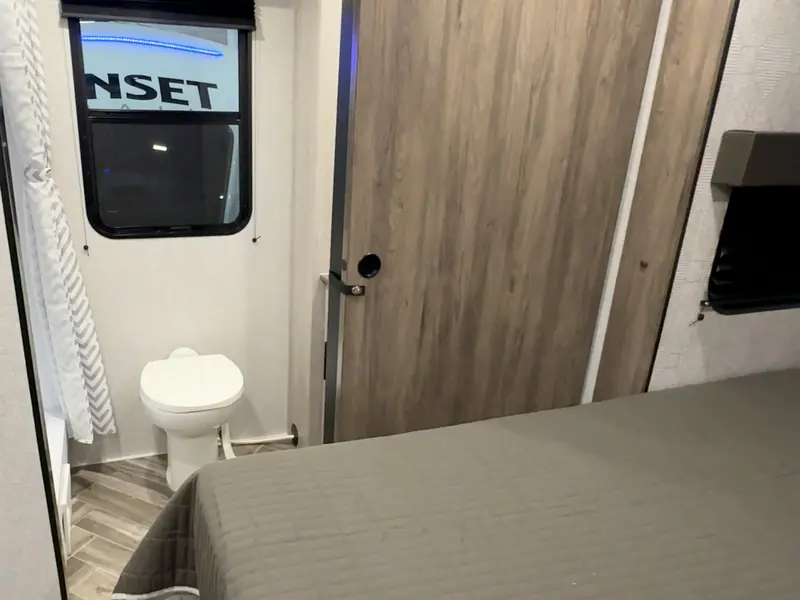 A Tecma electric toilet installed in the compact head of a small RV, seen through the open door beside the foot of the bed, with a hand-flush pedal and coiled discharge hose visible at its base