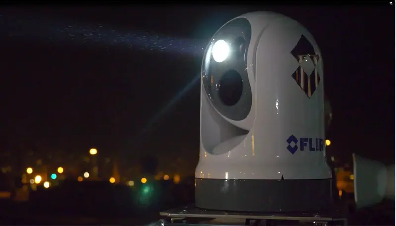 A FLIR pan and tilt marine thermal camera mounted on a mast at night, its lens dome lit from an integrated spotlight beside it, a city skyline glowing in the background