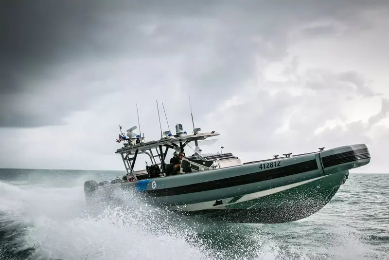 A law enforcement rigid inflatable boat launches off a wave in rough weather, a FLIR camera dome mounted on the radar arch above the console