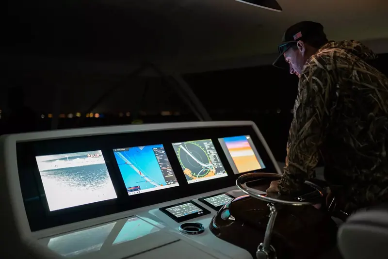 A boat captain in camouflage foul-weather gear stands at a night helm lit by a bank of navigation and thermal-camera displays