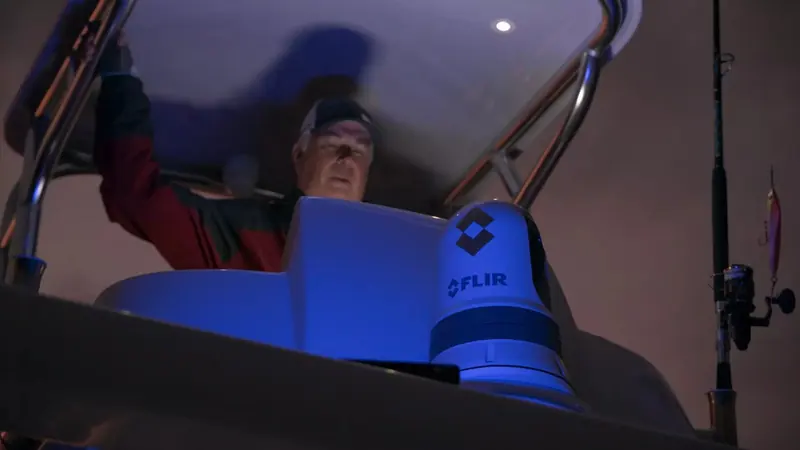 An angler at a night-fishing helm reaches up past a blue-lit FLIR thermal camera mounted on the hardtop, rod holders visible to the side
