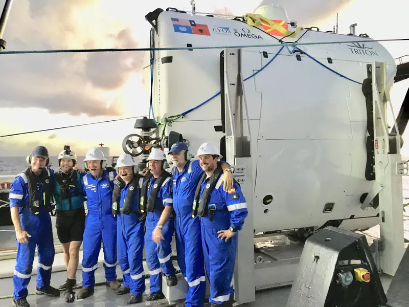 Pilot and support crew pose on deck beside Triton's full ocean depth submersible, its hull marked with EYOS Expeditions and OMEGA branding and the flags of the nations visited during the Five Deeps Expedition.