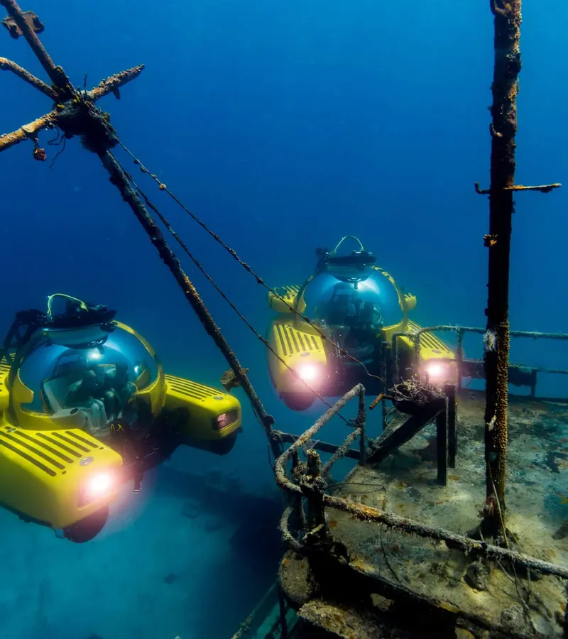 Two Triton submersibles moor beside the rigging of a sunken wreck, their acrylic spheres lit from within, during a wreck-diving deployment.