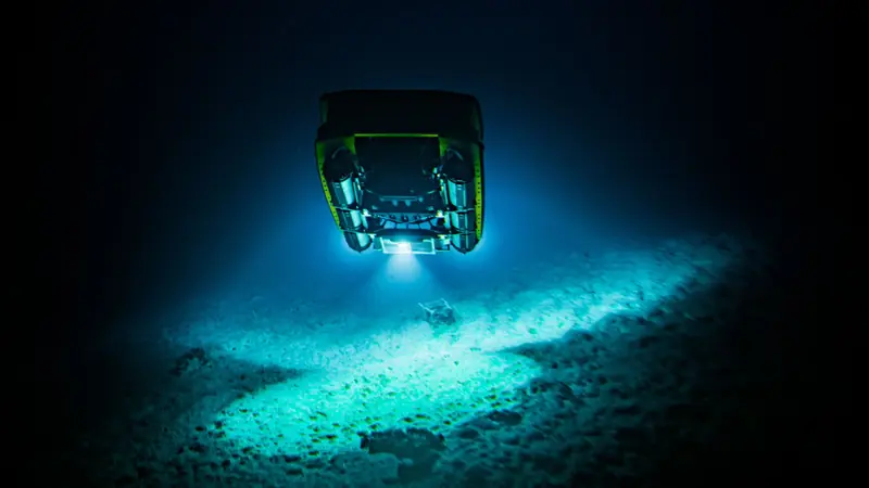 Seen from directly below, a Triton submersible's floodlight fans across bare seafloor, its ballast tanks and skid frame silhouetted against the water column.