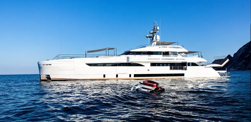 A submersible floats on the surface beside a white superyacht lying at anchor under a headland.