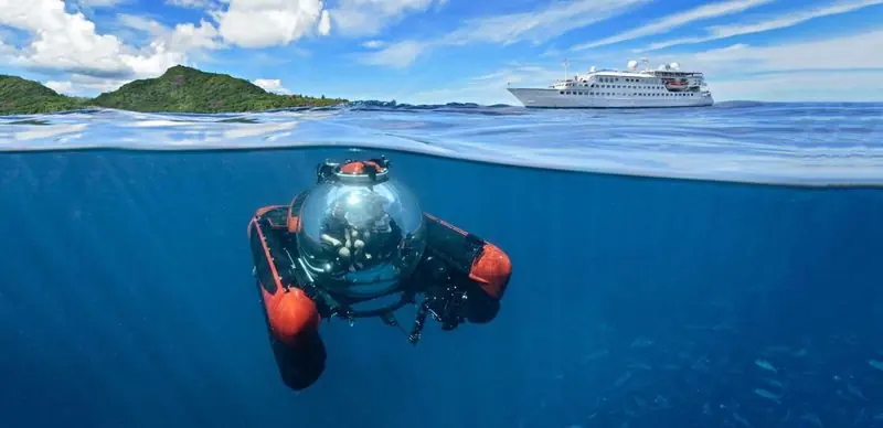 Split between air and water, a red and black submersible carries two people just below the surface while a ship sits off a green island.