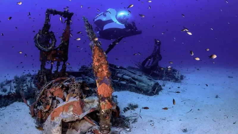 Deep over a sandy bottom, a submersible passes a scattered wreck with reef fish crowding the water.