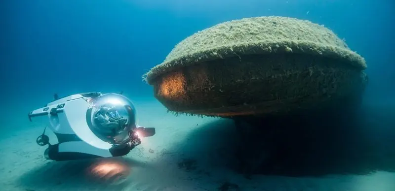Lights on, a white submersible hovers beside the weed-covered hull of a large wreck resting on the seabed.