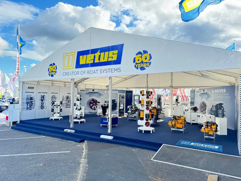 A white marquee marked VETUS, Creator of Boat Systems and 60 Years shelters rows of engines, thrusters and drive units on a navy blue show floor, flags flying against a blue sky outside.