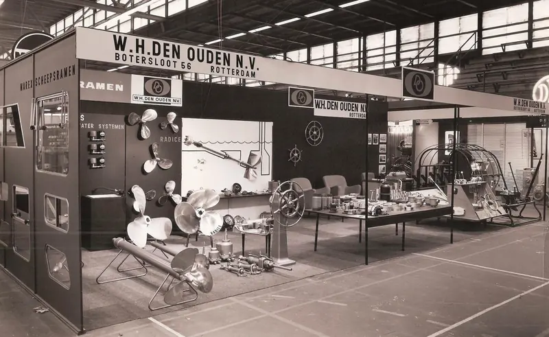 A 1960s trade show booth reads W.H. Den Ouden N.V., Botersloot 56, Rotterdam above racks of bronze propellers, a ship's wheel and steering gear laid out on tables.